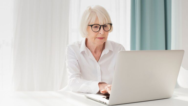 Person mit weißem Haar arbeitet an einem Laptop in einem hellen Raum mit weichem Fensterlicht und hellen, ruhigen Farbtönen.