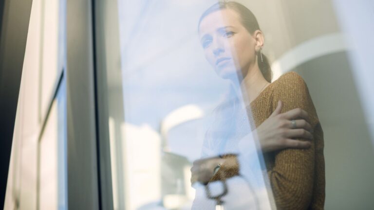 Person steht nachdenklich hinter einer Glasfront, die Arme leicht verschränkt, während weiches Licht und Reflexionen eine ruhige, introspektive Atmosphäre erzeugen.
