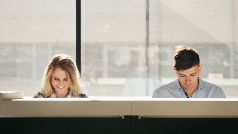 Zwei Personen sitzen nebeneinander an einem Tisch in einem hellen Raum und arbeiten konzentriert, während warmes Tageslicht durch ein großes Fenster fällt.