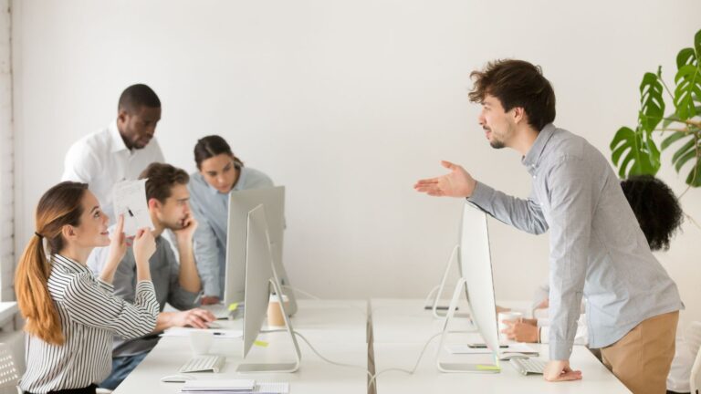 Mehrere Mitarbeitende arbeiten an Computern in einem modernen Büro, während eine Person über den Schreibtisch hinweg gestikulierend spricht