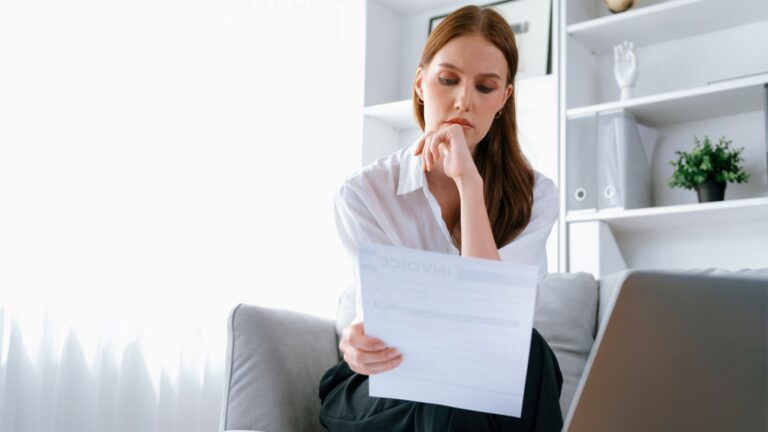Eine Frau sitzt in einem hellen, modernen Wohn‑ oder Arbeitsraum auf einem Sofa und liest aufmerksam ein Dokument.
