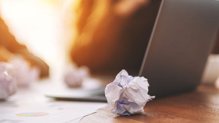 in zerknülltes Blatt Papier liegt im Vordergrund auf einem Holztisch. Im Hintergrund sind unscharf weitere Papierknäuel, ein aufgeklappter Laptop und Dokumente zu sehen, beleuchtet von warmem Licht. Stimmung der Trauer
