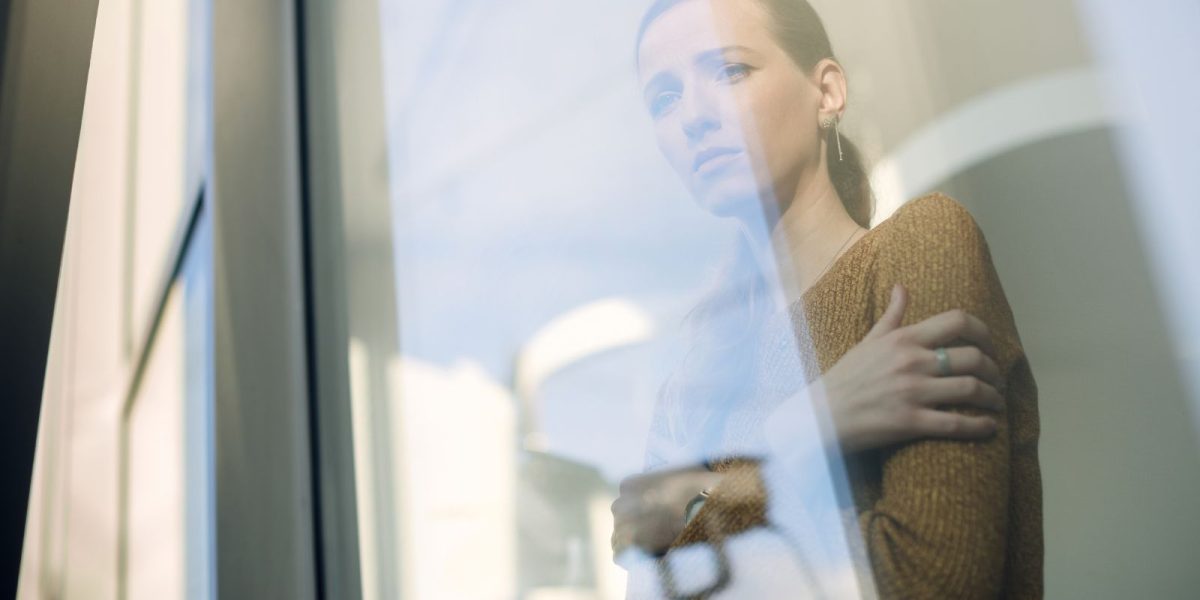 Person steht nachdenklich hinter einer Glasfront, die Arme leicht verschränkt, während weiches Licht und Reflexionen eine ruhige, introspektive Atmosphäre erzeugen.