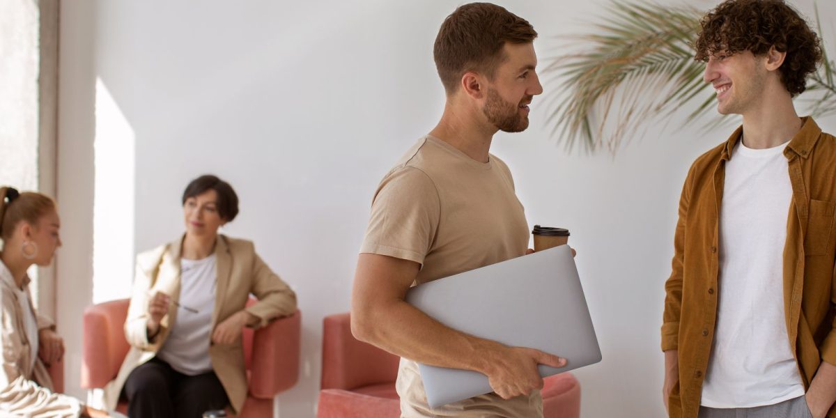 Mehrere Personen unterhalten sich in einer hellen Lounge, zwei stehen im Vordergrund mit Laptop und Getränken.