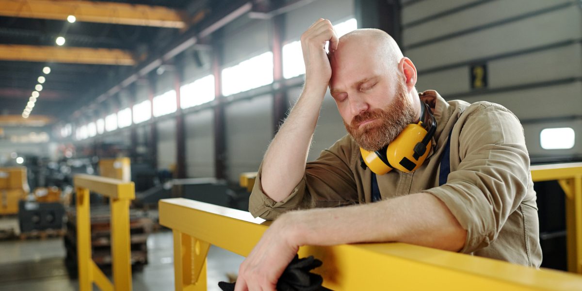 Tired or sick bald engineer touching his head while leaning by bar at break in the middle of working day in workshop