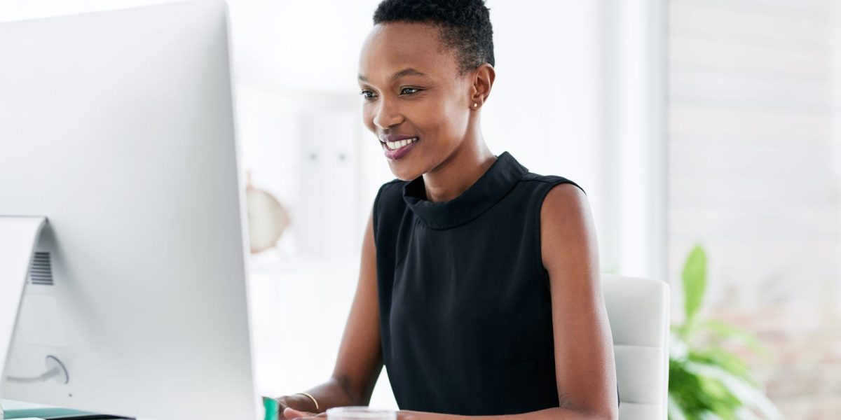 Women of Colour sitzt am Schreibtisch im Büro und schaut auf einen Computer und lächelt