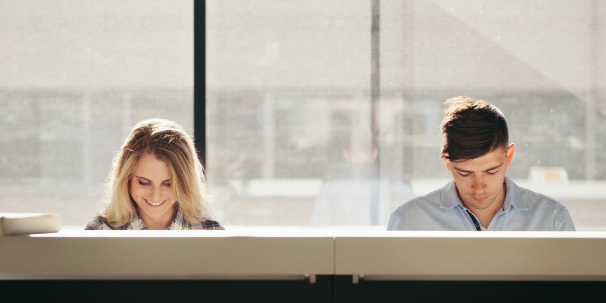 Zwei Personen sitzen nebeneinander an einem Tisch in einem hellen Raum und arbeiten konzentriert, während warmes Tageslicht durch ein großes Fenster fällt.