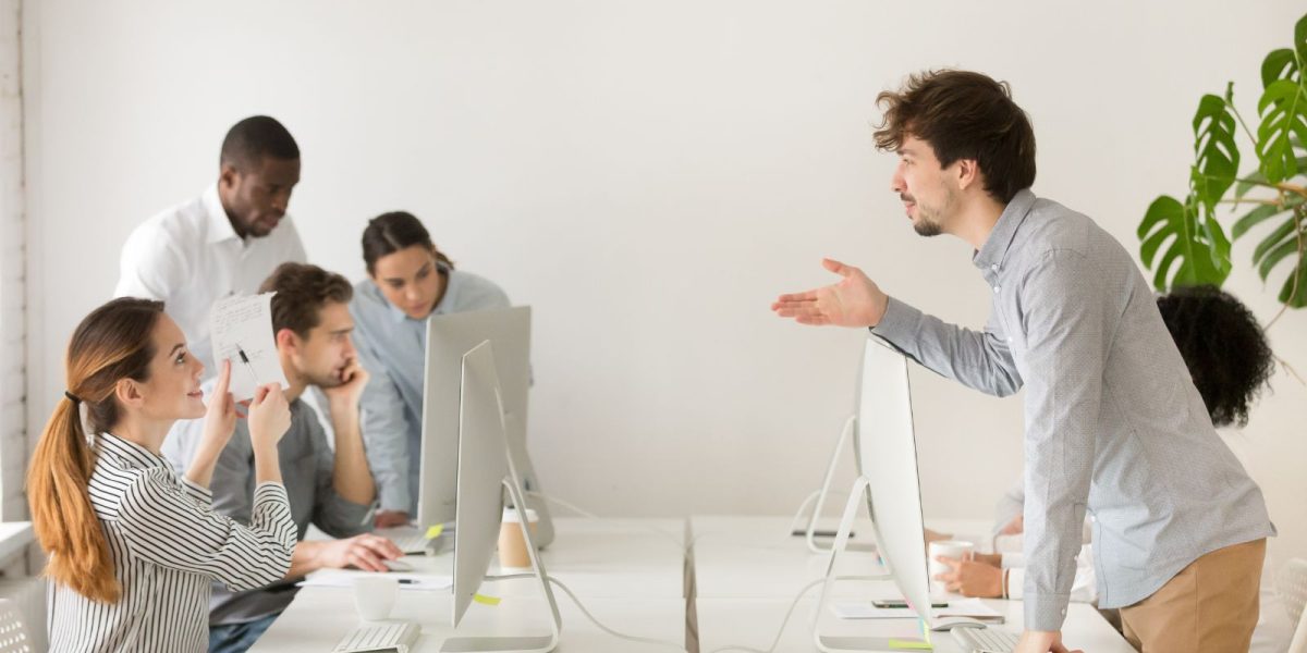 Mehrere Mitarbeitende arbeiten an Computern in einem modernen Büro, während eine Person über den Schreibtisch hinweg gestikulierend spricht