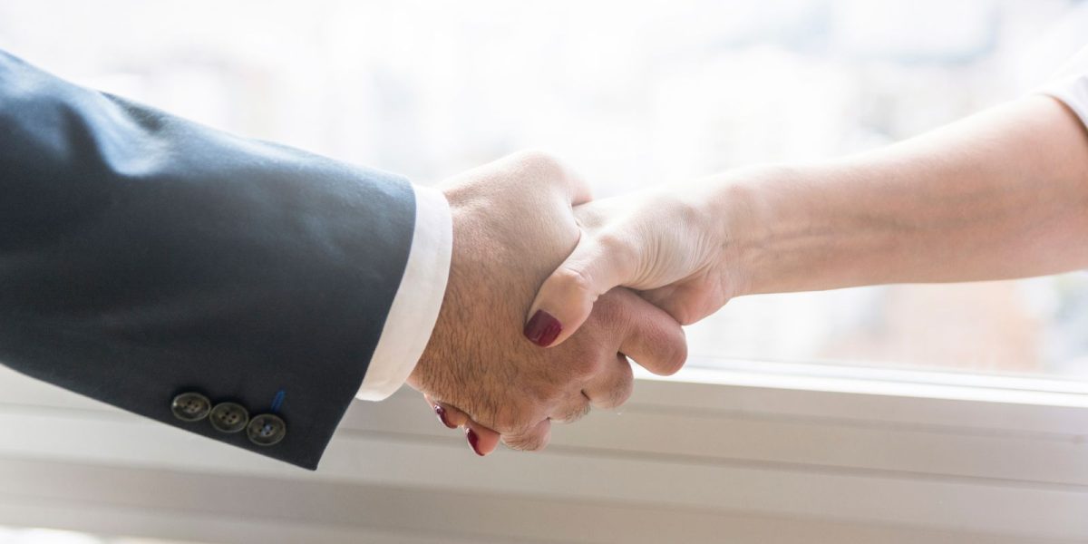 Zwei Hände, die sich vor einem hellen Fenster zu einem Handschlag treffen. Der Fokus liegt auf den Händen, während der Hintergrund unscharf ist.