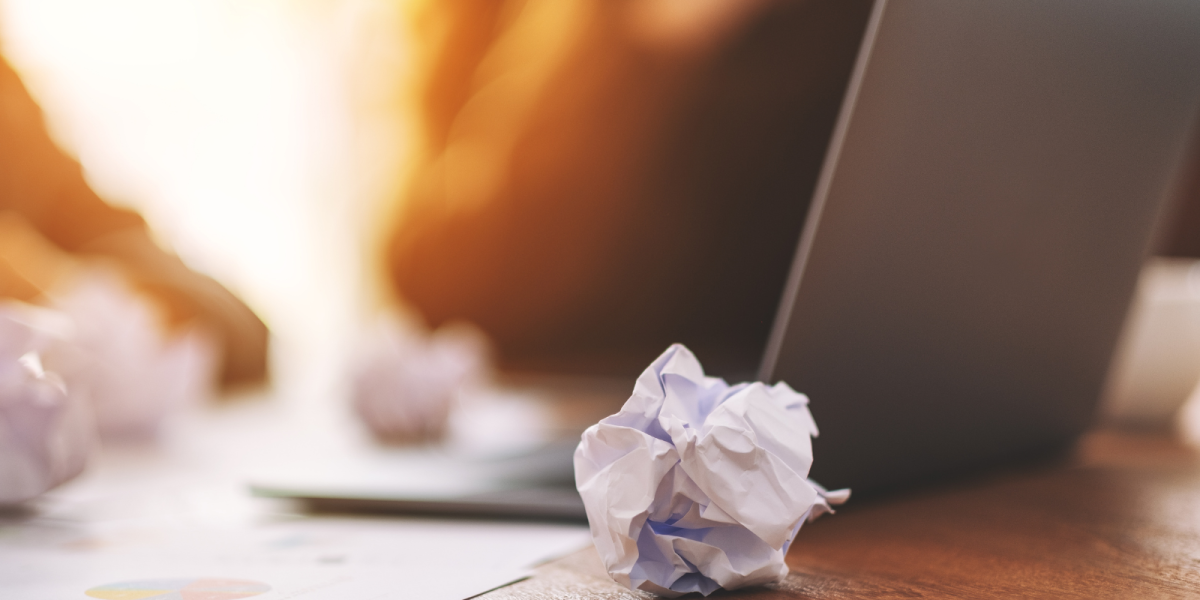in zerknülltes Blatt Papier liegt im Vordergrund auf einem Holztisch. Im Hintergrund sind unscharf weitere Papierknäuel, ein aufgeklappter Laptop und Dokumente zu sehen, beleuchtet von warmem Licht. Stimmung der Trauer