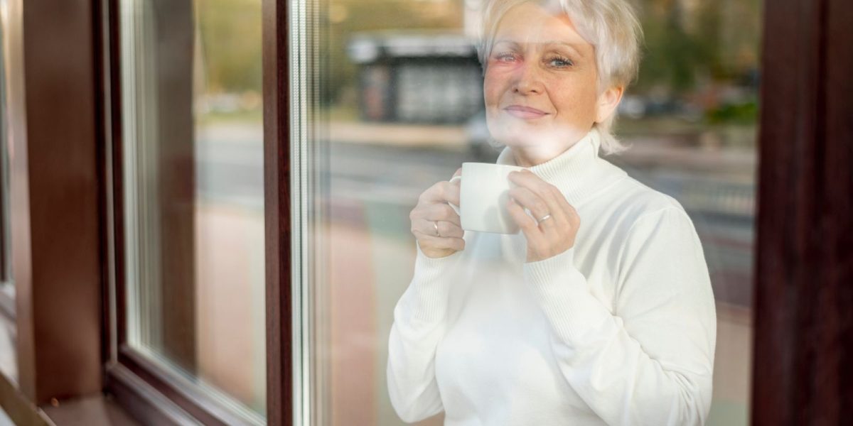 Ältere Frau steht mit Kaffeetasse am Fenster und schaut freundlich nachdenklich in die kamera