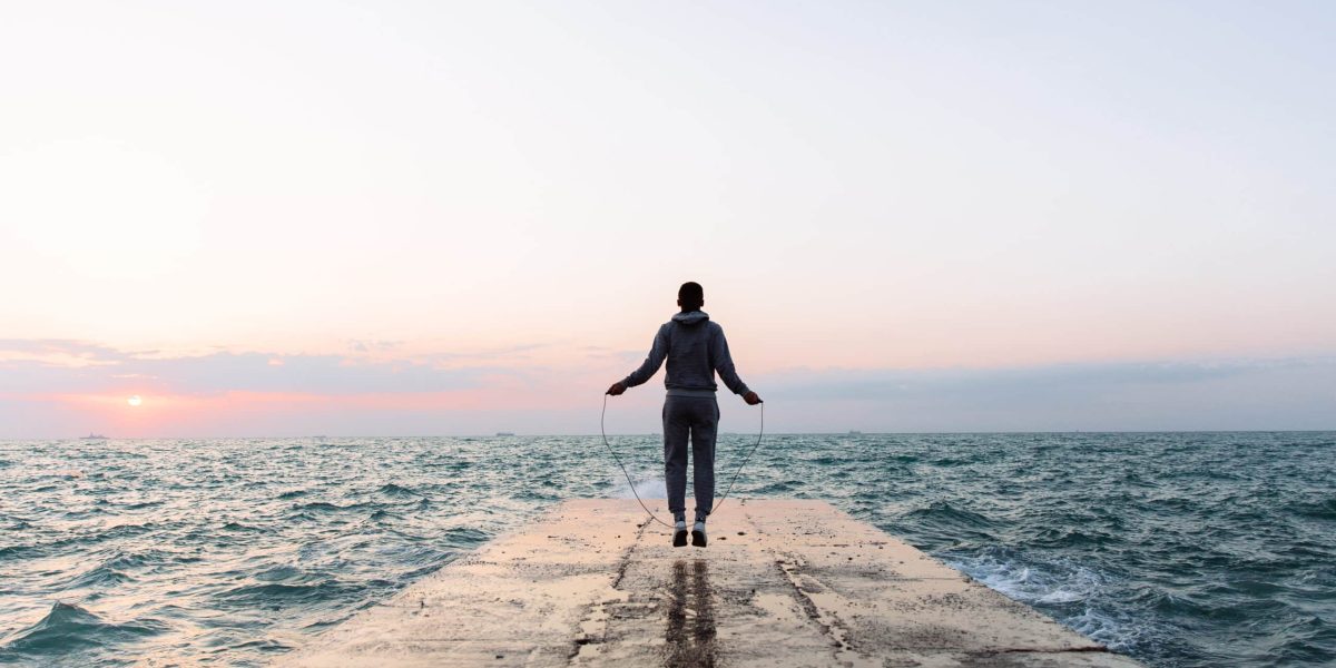 Full length view of young man in sportswear jumping with skipping rope, workout on pier, near the sea. View from back.