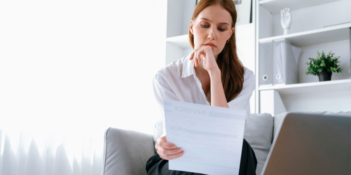 Eine Frau sitzt in einem hellen, modernen Wohn‑ oder Arbeitsraum auf einem Sofa und liest aufmerksam ein Dokument.